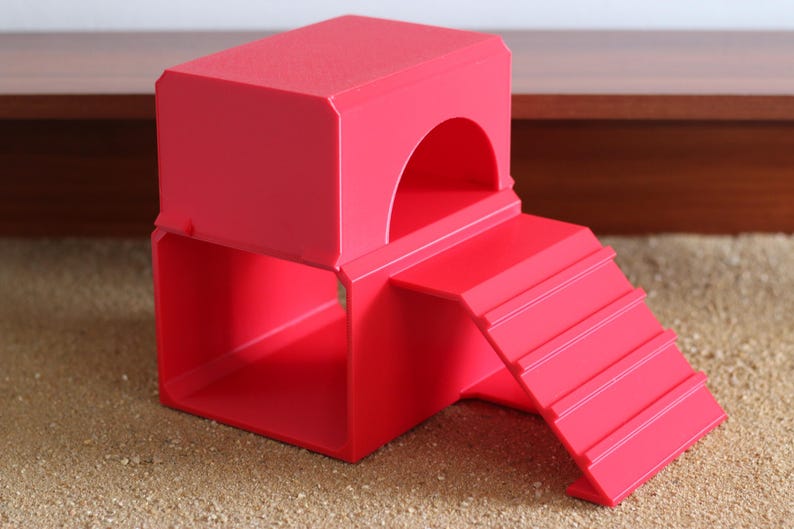 May include: A bright red plastic pet hideout featuring a two-level structure with a hollow square base and a top platform with an arched entrance. A set of stairs with four horizontal ridges is attached to the side, leading up to the top level.