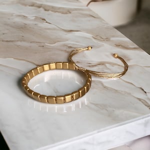 Peut inclure: Un bracelet en perles carrées dorées avec une fermeture en cordon doré. Le bracelet est posé sur une surface en marbre blanc.