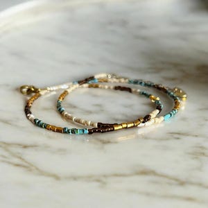 Puede incluir: Un delicado collar de capas con pequeñas cuentas blancas, marrones, doradas y turquesas.