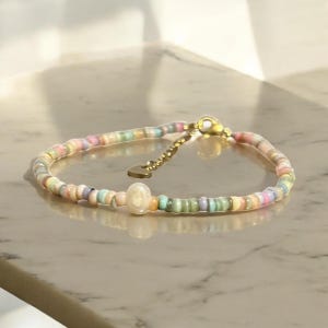 Peut inclure: Un bracelet délicat composé de petites perles aux couleurs pastel, dans des tons de rose, de jaune, de vert et de bleu. Le bracelet est doté d'un fermoir doré et d'un petit charme en forme de cœur. Une perle plus grande, blanc cassé, est centrée.