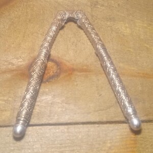 May include: Silver-colored nutcracker with ornate, textured handles. The nutcracker is open, displaying its V-shape. The handles have a detailed, repeating pattern. The tips of the handles are rounded and smooth.