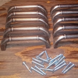 May include: A collection of antique-style cabinet pulls with a bronze finish, arranged in two stacks of five. The pulls have a ribbed center and curved ends. A pile of silver screws is also present.