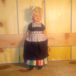 May include: A vintage doll dressed in traditional Dutch attire. The doll has blonde hair, blue eyes, and a white head covering. It wears a black dress with a patterned apron and a colorful striped skirt. The doll is standing in front of a wooden background.