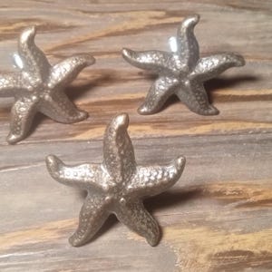 May include: Three silver-toned metal drawer knobs shaped like five-pointed starfish. Each knob features a textured, bumpy surface detail and a central mounting point on the back, arranged in a triangular formation on a flat surface.