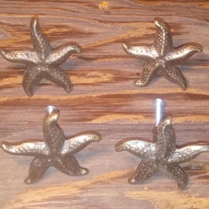 May include: Four bronze-colored starfish cabinet knobs. Each knob is a detailed starfish shape with five points. The knobs are mounted on a wooden surface, suggesting a coastal or nautical theme for home decor.