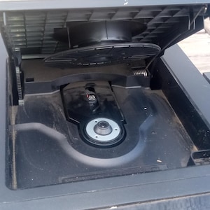 Peut inclure: Lecteur CD Sony noir avec le tiroir &agrave; disques ouvert. Le lecteur est noir avec le logo Sony argent&eacute;. Le tiroir &agrave; disques est ouvert, r&eacute;v&eacute;lant le m&eacute;canisme du disque. En arri&egrave;re-plan, un b&acirc;timent rouge et de l'herbe verte.