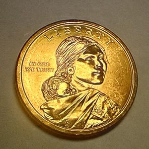 Puede incluir: Una moneda de un dólar de color dorado que presenta un perfil de Sacagawea, con la palabra "LIBERTY" en la parte superior y "IN GOD WE TRUST" inscrita. La moneda tiene un diseño detallado y un acabado brillante.