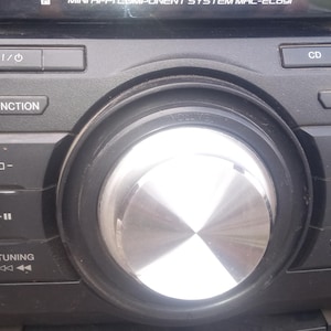 Peut inclure: Une mini-cha&icirc;ne st&eacute;r&eacute;o noire avec un gros bouton central argent&eacute; et divers boutons de fonction. Le syst&egrave;me comprend des boutons pour CD, iPod et commandes de r&eacute;glage. L'&eacute;cran affiche du texte en haut.