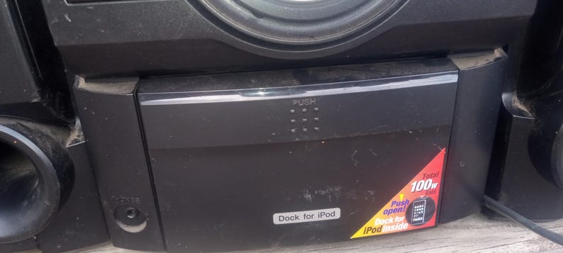 Peut inclure: Syst&egrave;me st&eacute;r&eacute;o noir avec un haut-parleur et une station d'accueil pour iPod. Le syst&egrave;me comporte un bouton "PUSH" et un autocollant indiquant "Dock for iPod" et "100w". Le syst&egrave;me a un cordon d'alimentation.