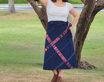 May include: A navy blue wrap skirt with white pinstripes and colorful woven accents. The skirt features a wrap design with a tie closure. The skirt is paired with a white short-sleeved top. The skirt is approximately knee-length.