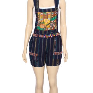 May include: Black and white striped short overalls with a colorful embroidered patch on the front. The overalls have a thin, colorful belt.