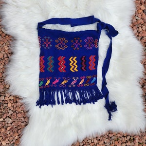 May include: A blue embroidered fabric with colorful geometric patterns and fringe. The fabric has a tie at the top.