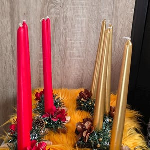 May include: Four taper candles, two red and two gold, each standing in a star-shaped wooden base. The bases are decorated with green tinsel, small gold beads, and red or brown floral accents. The candles are arranged in a group on a bright orange faux fur surface.