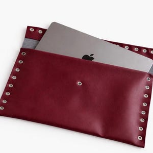 Puede incluir: Una funda para portátil burdeos con ojales plateados en los bordes. Un portátil plateado con el logotipo negro de Apple es parcialmente visible en el interior. La funda está hecha de un material liso, similar al cuero.