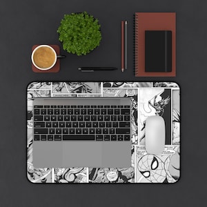 May include: A black and white comic book-themed desk mat featuring panels with various superhero characters. A laptop, white mouse, coffee cup, plant, pen, pencils, and notebooks are on the dark surface.