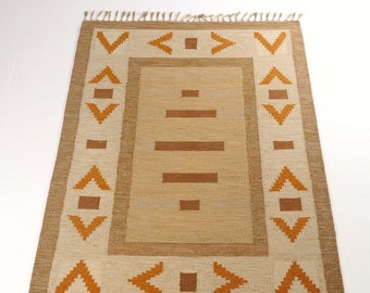 May include: Rectangular beige rug with a geometric design. The rug features a central beige field with brown horizontal lines and squares. The border is beige with brown geometric shapes and fringe on the short ends.