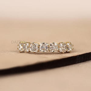 May include: A gold band ring featuring five square-cut, clear gemstones. The ring is set on a neutral-toned surface, with the brand name "DAZZIO" visible in the background. The gemstones are evenly spaced and reflect light.