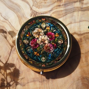 K&ouml;nnte beinhalten: Ein rundes, goldfarbenes Kompakt mit einem floralen Design. Das Design zeigt einen Strau&szlig; aus rosa, wei&szlig;en und gelben Blumen mit gr&uuml;nen Bl&auml;ttern auf blauem Hintergrund. Das Kompakt hat einen Klappdeckel.