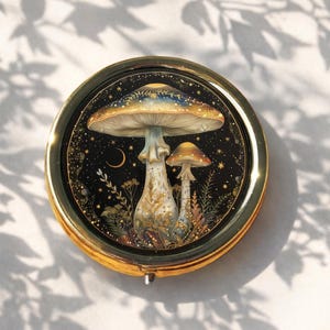 May include: A circular, gold-coloured compact mirror with a mushroom illustration. The artwork shows two mushrooms set against a starry night sky with a crescent moon. The compact is on a white surface with leaf shadows.