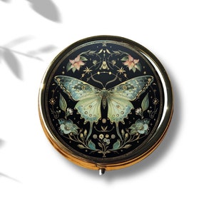 May include: A round, gold-rimmed compact mirror with a black background and a detailed illustration of a butterfly with floral accents. The butterfly has green and blue wings with gold accents.