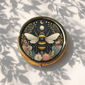May include: A circular compact mirror with a gold frame, showcasing a bee design. The bee is central, with floral and moon phase details on a dark backdrop. The compact sits on a white surface, with shadows of leaves cast across it.