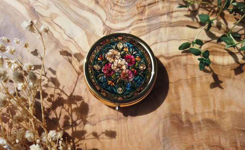 K&ouml;nnte beinhalten: Ein rundes, goldfarbenes Kompakt mit einem floralen Design. Das Design zeigt einen Strau&szlig; aus rosa, wei&szlig;en und gelben Blumen mit gr&uuml;nen Bl&auml;ttern auf blauem Hintergrund. Das Kompakt hat einen Klappdeckel.