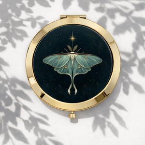 May include: A circular, gold-coloured compact mirror featuring a detailed illustration of a luna moth on a dark blue background. The moth is light green with delicate wing patterns. A star and leaves are above the moth. The compact is open.