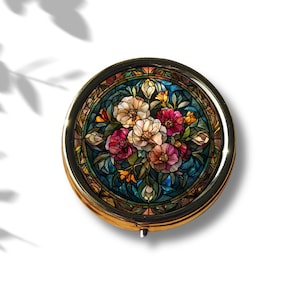 K&ouml;nnte beinhalten: Eine runde, goldfarbene Schmuckdose mit einem Buntglasdesign, das einen Strau&szlig; aus rosa, wei&szlig;en und roten Blumen auf blauem Hintergrund zeigt.