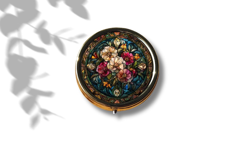 K&ouml;nnte beinhalten: Eine runde, goldfarbene Schmuckdose mit einem Buntglasdesign, das einen Strau&szlig; aus rosa, wei&szlig;en und roten Blumen auf blauem Hintergrund zeigt.