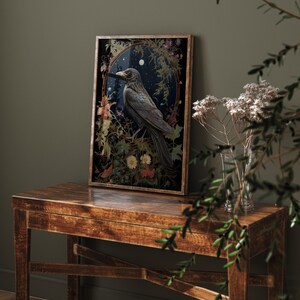 May include: A framed print of a black raven perched on a branch in a forest setting. The raven is surrounded by flowers and leaves, and the background is a dark blue sky with stars. The print is in a gold frame.