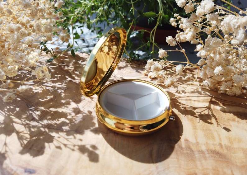 K&ouml;nnte beinhalten: Eine goldene Pillendose mit einem wei&szlig;en Innenraum, der in drei Abschnitte unterteilt ist. Die Dose ist ge&ouml;ffnet und der Deckel liegt auf der Seite.