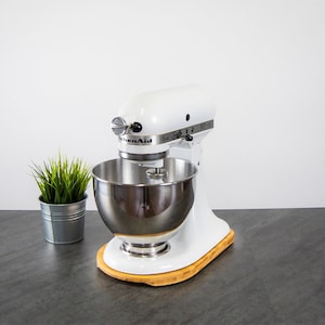 Pode incluir: Batedeira de pedestal KitchenAid branca com uma tigela de aço inoxidável e uma base de madeira. A batedeira está em uma bancada cinza.