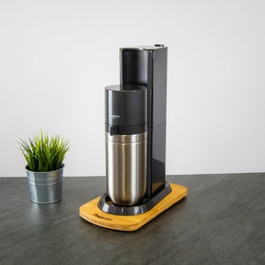 Könnte beinhalten: Ein schwarzer und silberner Sodamacher mit einer Edelstahlflasche und einem Bambussockel.