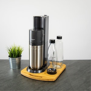 Könnte beinhalten: Eine schwarze und silberne SodaStream-Maschine mit einem Bambussockel. Die Maschine hat zwei klare Glasflaschen mit schwarzen Kappen. Eine Flasche trägt den Schriftzug "SodaStream".