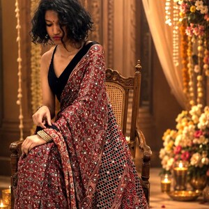 May include: A deep red and black saree featuring intricate geometric patterns and small circular mirror embellishments throughout the fabric. The garment is paired with a sleeveless black blouse with a V-neckline, creating a cohesive ensemble.