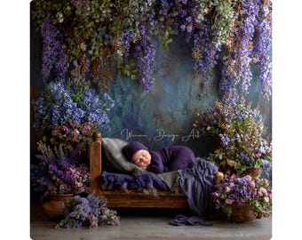 May include: A newborn baby in a purple outfit and hat sleeps peacefully on a small wooden bed. The bed is draped with a gray pillow and a purple blanket. The background features a floral arrangement with purple and blue flowers.