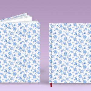 May include: Two white notebooks with a blue floral pattern. The notebook on the left is open, showing blank pages. The notebook on the right is closed and has a red ribbon bookmark.