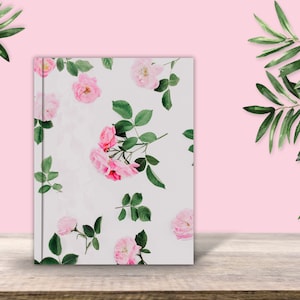 May include: A white notebook with a floral design featuring pink roses and green leaves. The notebook is on a wooden surface.