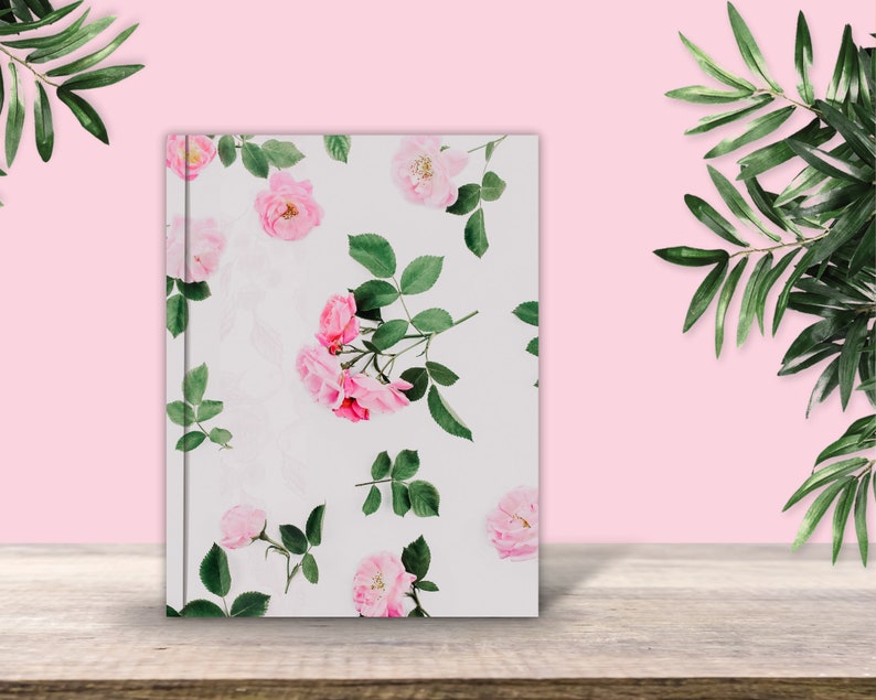 May include: A white notebook with a floral design featuring pink roses and green leaves. The notebook is on a wooden surface.