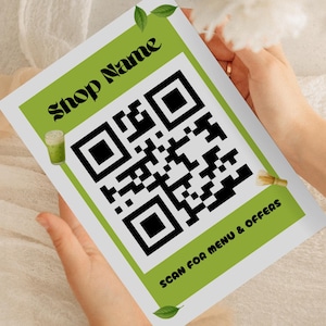 May include: A white and green printed card with the text "Shop Name" and a large QR code. The card also includes the text "SCAN FOR MENU & OFFERS". The card is held by a person.