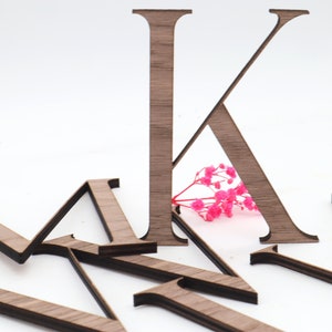 Könnte beinhalten: Holzbuchstabe K mit brauner Holzmaserung. Der Buchstabe steht aufrecht auf einer weißen Oberfläche. Im Hintergrund befinden sich weitere Holzbuchstaben.