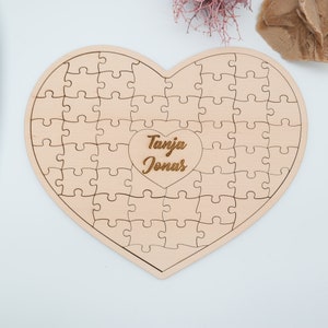 May include: A wooden heart-shaped jigsaw puzzle with the names "Tanja Jonas" engraved in the centre. The puzzle is made of light-coloured wood and has a smooth finish.