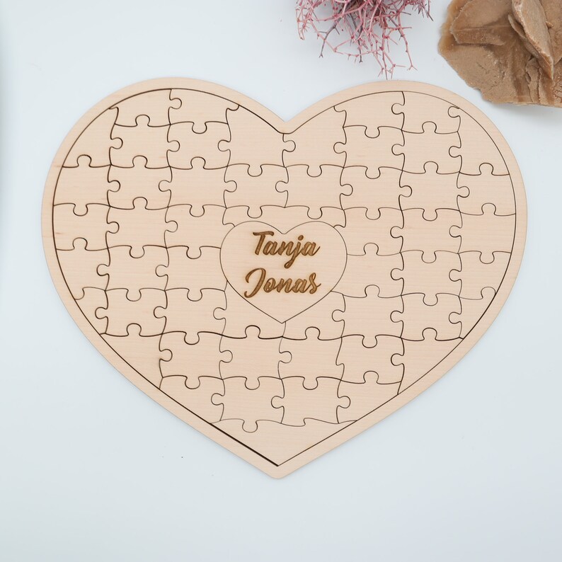 May include: A wooden heart-shaped jigsaw puzzle with the names "Tanja Jonas" engraved in the centre. The puzzle is made of light-coloured wood and has a smooth finish.