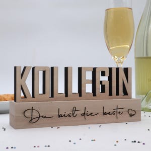 Könnte beinhalten: Ein Holzschild mit dem Wort "KOLLEGIN" in großen, ausgeschnittenen Buchstaben. Darunter steht in Schreibschrift "Du bist die beste". Ein Glas Sekt und eine Weinflasche sind im Hintergrund.
