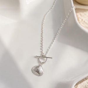 May include: A silver-toned necklace featuring a delicate cable chain and a toggle clasp closure. A small, textured seashell charm hangs from the circular ring of the toggle, creating a simple and cohesive metallic design with a polished finish.