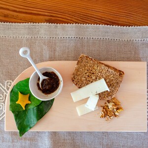 Puede incluir: Una tabla de cortar de madera con una rebanada de pan, queso, nueces y un pequeño tazón de mermelada. La tabla está sobre un mantel beige.