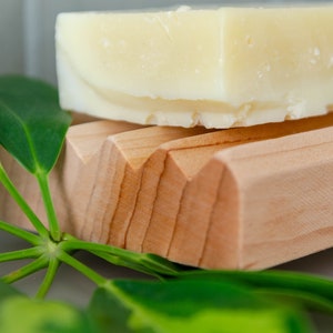Puede incluir: Una barra de jabón blanca descansando sobre un jabonera de madera con veta de madera natural. La jabonera tiene un borde ondulado. Hay hojas verdes en primer plano.