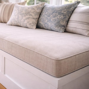 May include: A window seat with a beige cushion and patterned pillows in shades of beige and blue. The base of the seat is white, and the floor is wooden. The pillows add a decorative touch to the seating area.