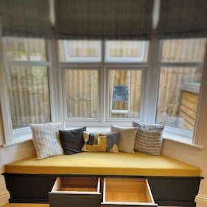 Op de afbeelding: Een vensterbank met een mosterdgeel kussen en diverse sierkussens. De bank heeft twee open lades eronder. Het raam heeft een patroon en kijkt uit op een houten hek.