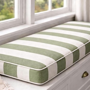 Puede incluir: Un cojín rectangular para asiento de ventana con anchas rayas alternas en verde oliva y blanco. El cojín tiene un borde ribeteado blanco y está colocado sobre un asiento de ventana blanco. La ventana es visible en el fondo.